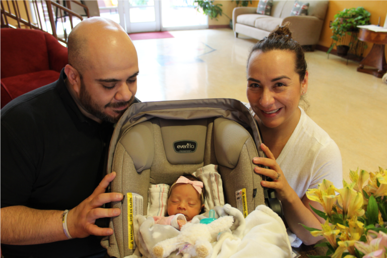 Newborn Carpena sits in her carseat while her parents are smiling towards camera